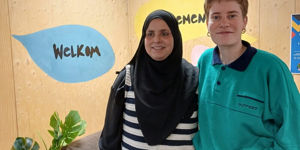 Mel en Nasira, opbouwwerkers van SAAMO Brussel poseren arm in arm voor de tekst "welkom"