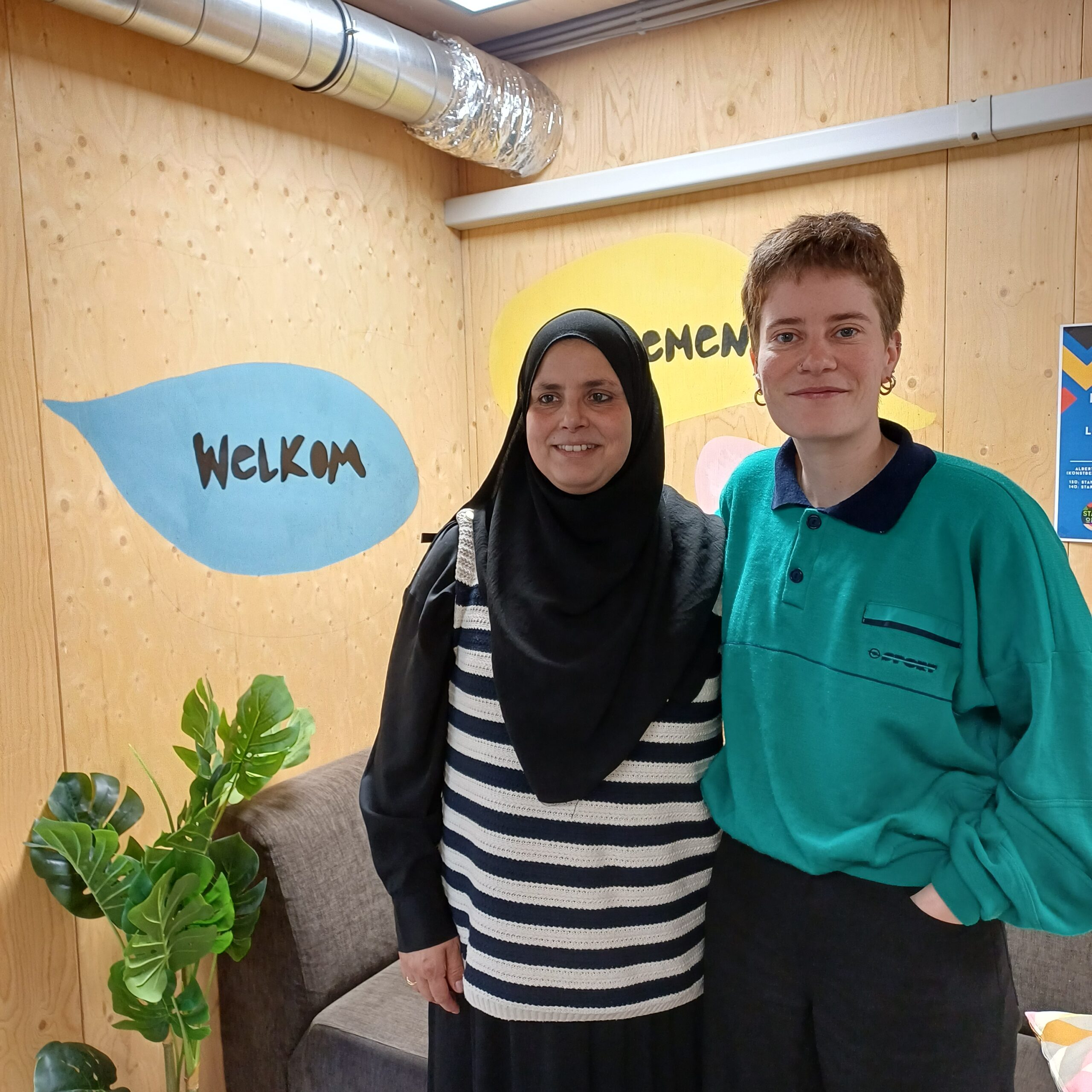 Mel en Nasira, opbouwwerkers van SAAMO Brussel poseren arm in arm voor de tekst "welkom"